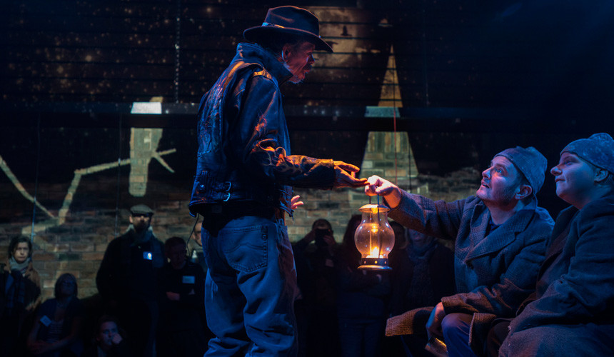 Jez Colborne in a leather jacket and cowboy hat holds a lamp up to a man and a woman in scruffy hobo costumes