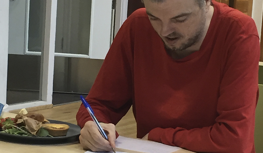 A man wearing a red jumper is drawing on the pages of the doodle book next to a plate of food.